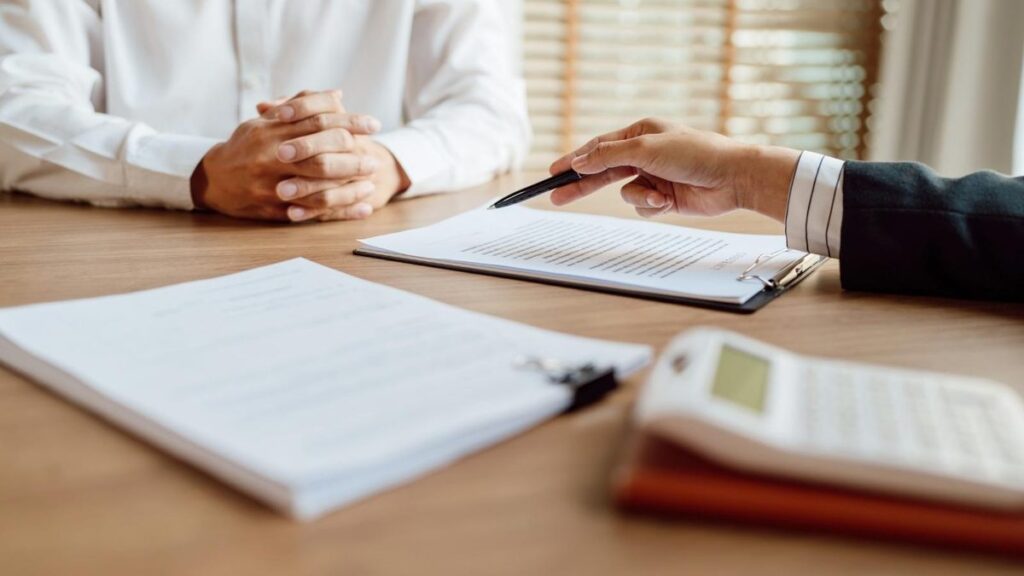 An adjuster points a pen at paperwork on a desk while a person sits across the table with a calculator nearby.