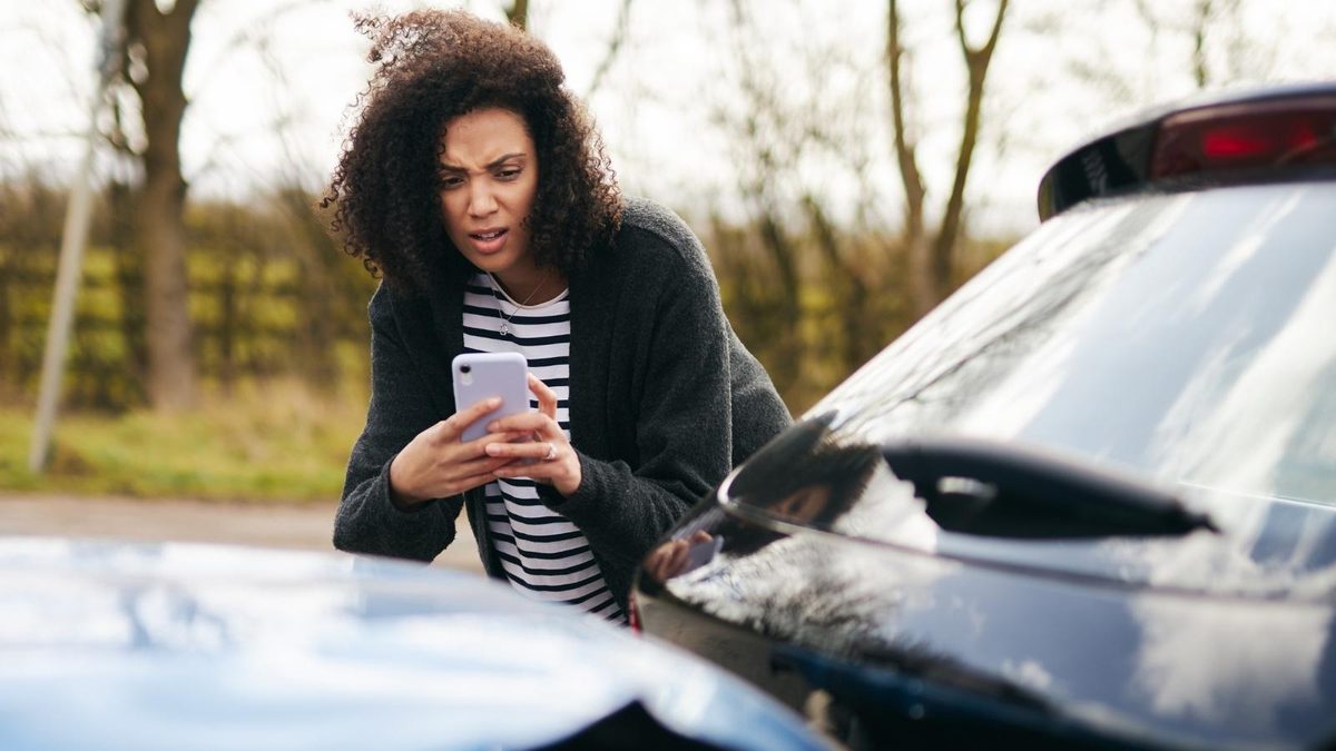A woman leans over her damaged car after a crash, looking at her phone with a worried expression.